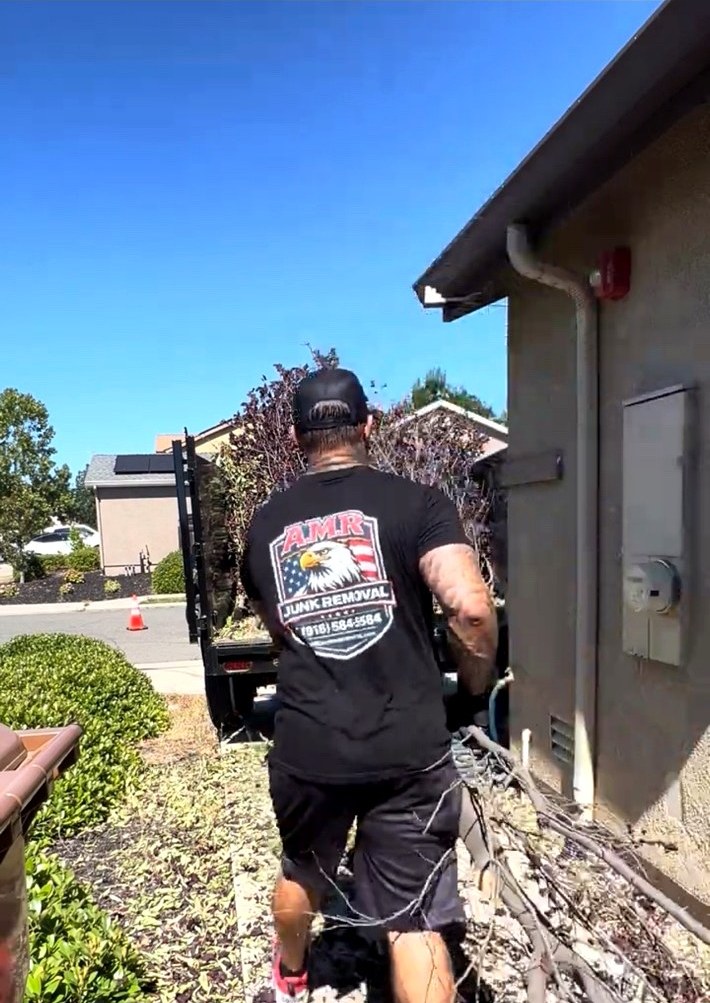 AMR Junk Removal team member in a branded company shirt carrying branches out of a residential side yard