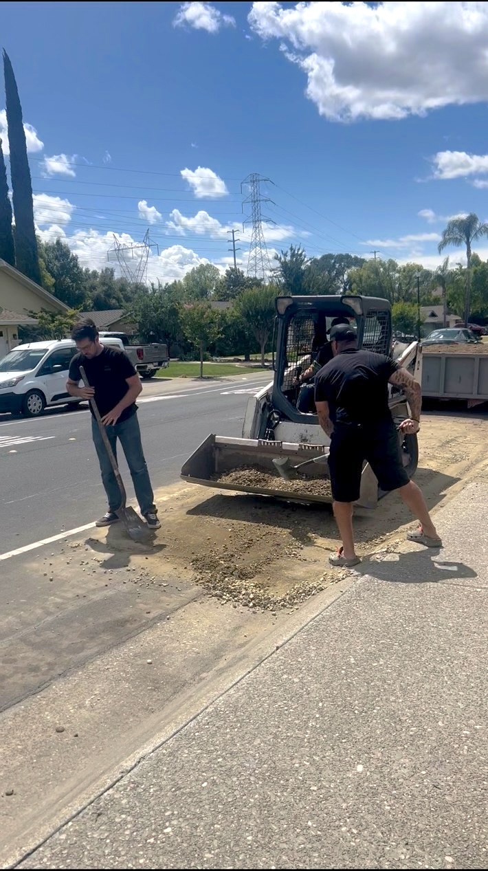 Two AMR Junk Removal crew members clearing gravel and debris at a curb with a skid steer and shovels